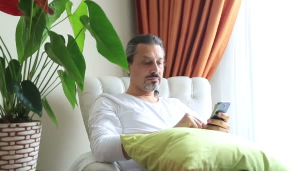 Hombre de mediana edad usando teléfono móvil. Relajado y sentado en el sofá interior — Vídeos de Stock