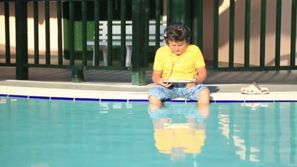 Νεαρό αγόρι με το ακουστικό με ψηφιακή δισκίο κοντά στην πισίνα — Αρχείο Βίντεο