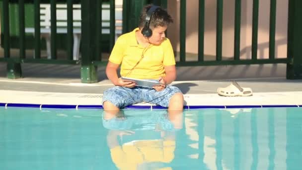 Jeune garçon avec écouteur utilisant une tablette numérique près de la piscine — Video