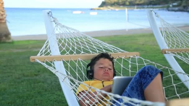 Niño con auriculares usando almohadilla i en una hamaca — Vídeo de stock