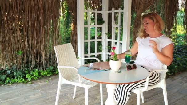 Женщина сидит за столом в летнем кафе с телефоном и ждет кого-то — стоковое видео