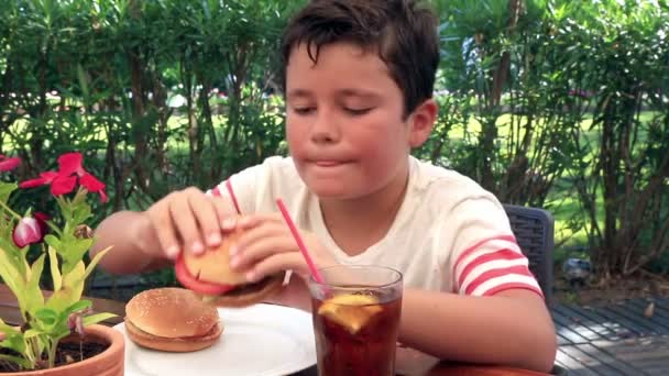Голодний хлопчик їсть гамбургер у кафе — стокове відео