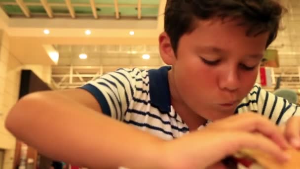 Głodny młody chłopak jedzenia hamburgera w restauracji — Wideo stockowe