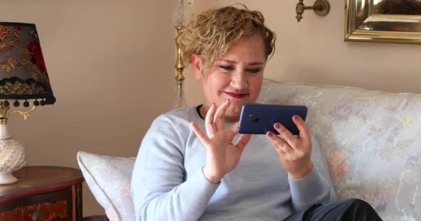中年女性座っていますソファと使用スマートフォンで自宅 動画クリップ