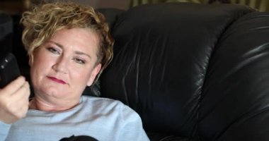 Uzaktan kumandalı mutlu orta yaşlı kadın evdeki kanepede oturmuş televizyon izliyor.