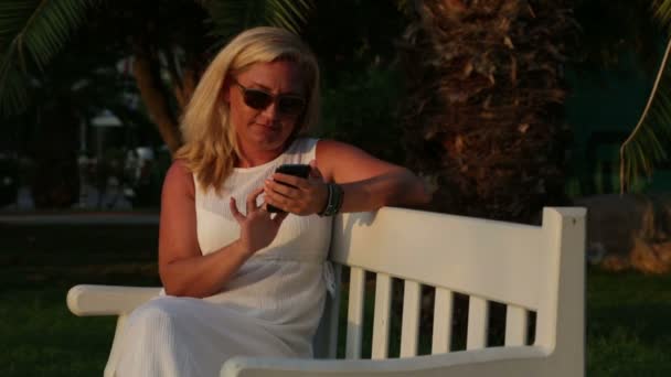 Женщина сидит на скамейке в парке со своим телефоном — стоковое видео