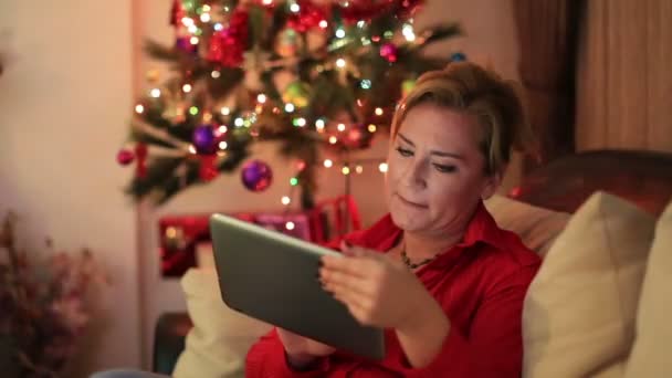 Portréja egy mid felnőtt nő ül a kanapé-közel a karácsonyfa, digitális tabletta — Stock videók