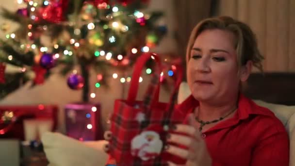 Портрет удивленной женщины с подарочной коробкой, сидящей на диване возле рождественской елки — стоковое видео