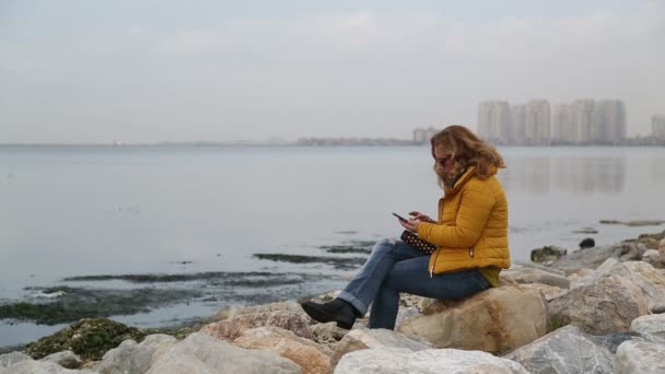 Жінка сидить на березі моря і використовує смартфон — стокове відео