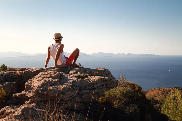 Contemplative young man sitting on the top of the mountain looking at distance at sunset with the sea in front