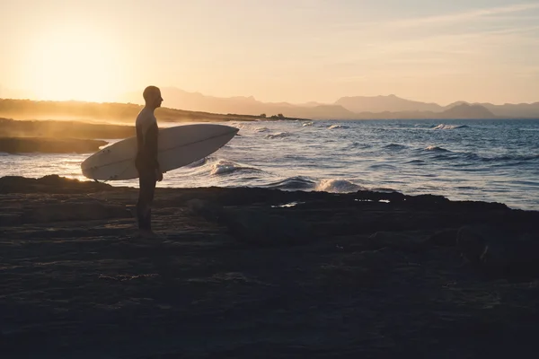 Jeune surfeur tenant une planche de surf contemplant les vagues au coucher du soleil Image En Vente