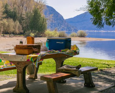 Gölün ve dağların arkasındaki parkta yapılacak piknik için kutular dolusu yiyecek ve tahta masa. Harrison Gölü, BC, Kanada. 15 Nisan 2021. Sokak manzarası, seyahat fotoğrafı, kimse yok..
