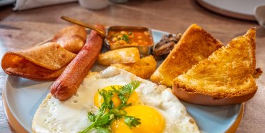 Kızarmış yumurta sarısı, ızgara sosis, kızarmış pastırma ve tost ile sağlıklı geleneksel İngiliz kahvaltısı. Güneşli tarafı yumurta ve sosis ile ahşap masada bir tabakta. Kapat.