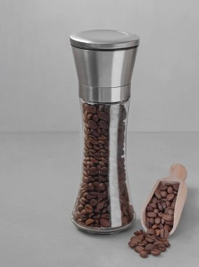 Gri arka planda gümüş kapaklı uzun cam bir kavanozda kahve çekirdekleri. Uzayı kopyala Dikey oryantasyon. Teneke kutunun tepesine odaklan. Yakınlarda kahve çekirdekli tahta bir kaşık var..