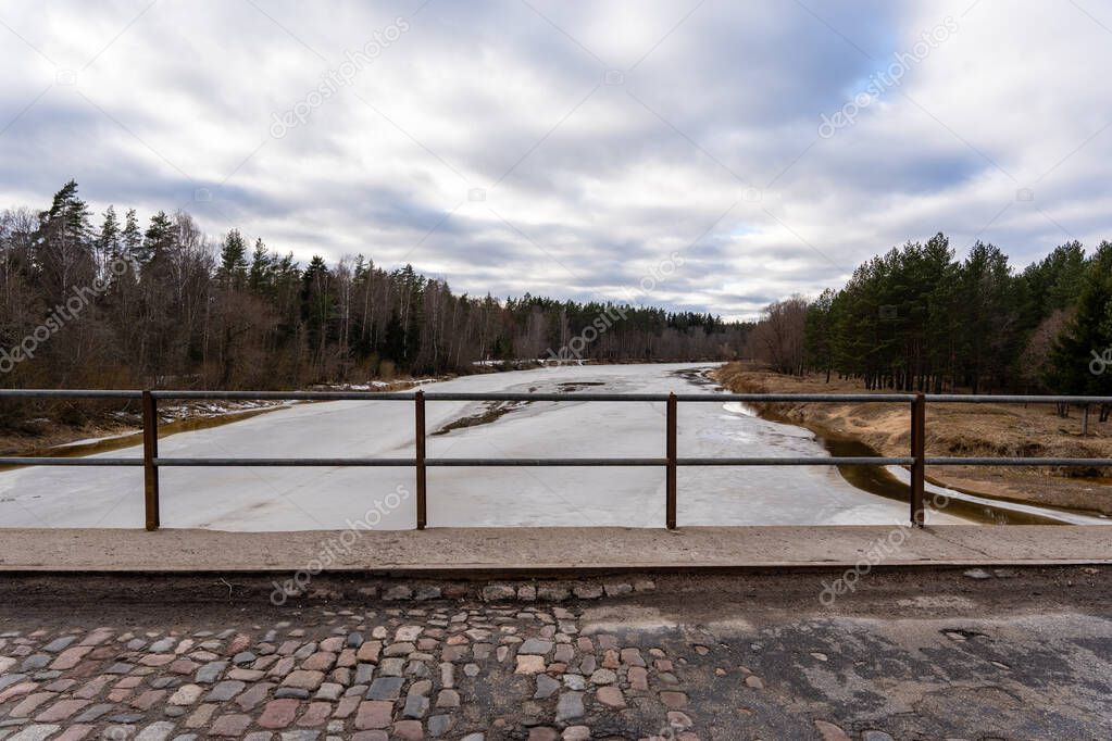 The longest river in Latvia is the Gauja, in the spring when the snow has melted and there is brown old grass along the edges but there is still ice on the river, the bridge railings are in the foreground.