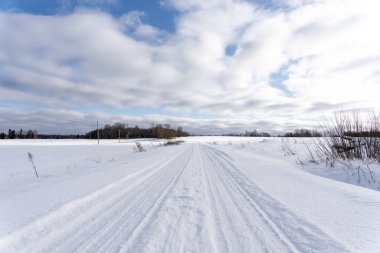 a beautiful sunny winter day in the Latvian countryside with blue skies and white clouds where there is a road in the middle of the field that is covered with snow and on which the tracks of car tires remain in the snow