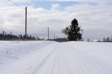 a beautiful sunny winter day in the Latvian countryside with blue skies and white clouds where there is a road in the middle of the field that is covered with snow and on which the tracks of car tires remain in the snow
