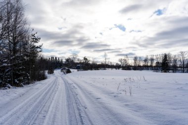 a beautiful sunny winter day in the Latvian countryside with blue skies and white clouds where there is a road in the middle of the field that is covered with snow and on which the tracks of car tires remain in the snow