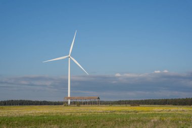 In the spring, a large rape field with a forest and blue skies behind it, there is a wind generator with a large propeller in the middle