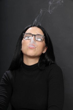 Siyah saçlı, gözlüklü, stüdyoda poz veren ve arka planda puro içen çekici Avrupalı bir kız. Tarz, trendler, sigara konsepti