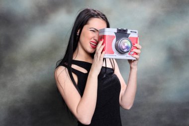 Uzun siyah saçlı güzel Avrupalı kız. Stüdyoda tuvalin arkasında poz veriyor ve elinde sahte bir kamera var. Tarz, trendler, moda konsepti.