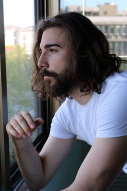 Uzun saçlı ve sakallı şık, beyaz bir adam balkonda poz veriyor. Tarz, trendler, moda konsepti.