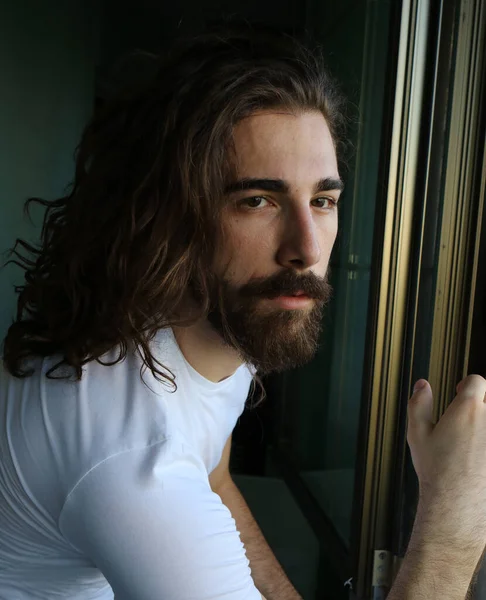 Stylish young caucasian guy with long hair and beard is posing on the balcony. Style, trends, fashion concept.