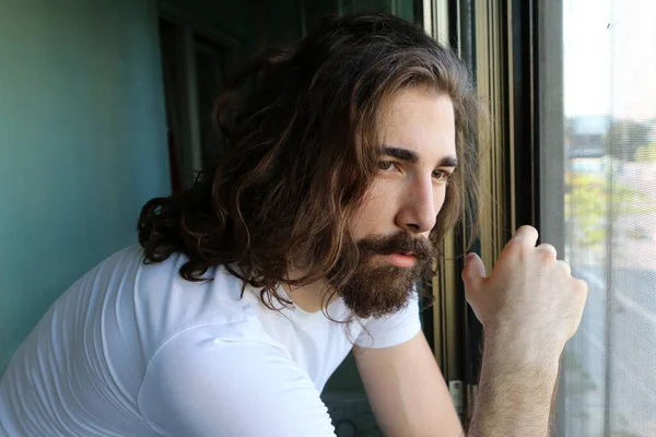 Stylish young caucasian guy with long hair and beard is posing on the balcony. Style, trends, fashion concept.