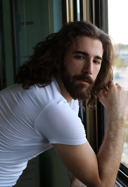 Stylish young caucasian guy with long hair and beard is posing on the balcony. Style, trends, fashion concept.