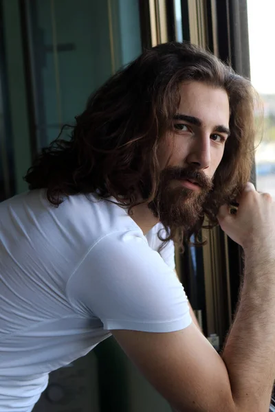 Stylish young caucasian guy with long hair and beard is posing on the balcony. Style, trends, fashion concept.