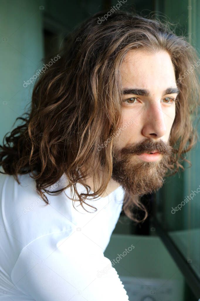 Stylish young caucasian guy with long hair and beard is posing on the balcony. Style, trends, fashion concept.
