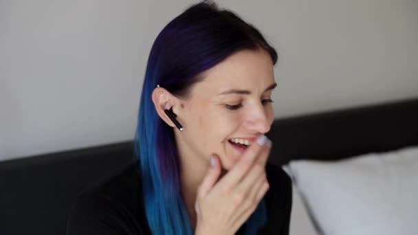Femeia tânără cu păr albastru râde așezată pe pat purtând căști fără fir — Videoclip de stoc