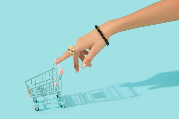 Beautiful groomed womans hand with fashionable ring holding shopping cart on blue background — Stock Photo, Image