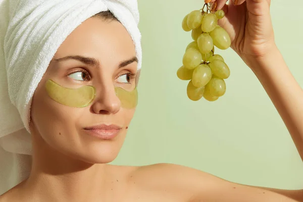 Mujer que usa parches para los ojos a base de uva sobre fondo verde pastel. Cuidado natural sostenible de la piel — Foto de Stock