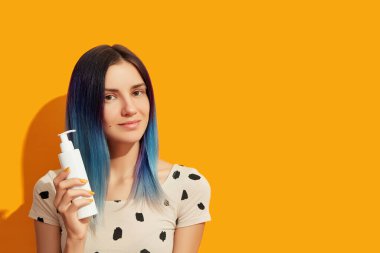 Genç, güzel bir kadın, mavi saçlı, beyaz kozmetik şişesini parlak arka planda tutuyor.