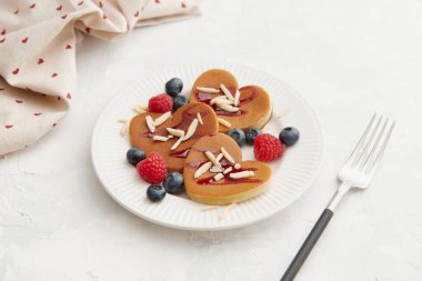 İçinde ahududu, yaban mersini, badem gevreği ve kırmızı böğürtlen sosu olan bir yığın kalp şeklinde krep ve gri arka planda beyaz tabakta çatal ve kalp baskılı doku. Sevgililer Günü konsepti 