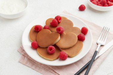 Beyaz tabakta bir yığın kalp şeklinde krep ahududu, çatal bıçaklı yoğurt ve beyaz desenli arka planda pembe peçete dokusu. Sevgililer Günü konsepti 