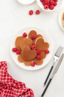 Beyaz tabakta ahududu, yoğurt, çatal bıçak takımı ve beyaz desenli arka planda kırmızı kareli peçete dokusu olan bir yığın kalp şeklinde krep. Sevgililer Günü konsepti 