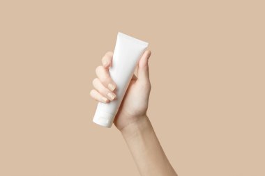 A womans hand holding a white cosmetic tube on a beige background. Care products beauty treatment concept