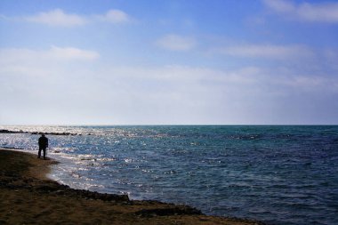 Sopalı yaşlı bir adamın silueti, sahil boyunca denize bakıyor. Uzakta, denizin kenarında tek başına.