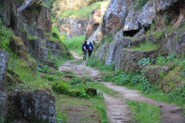 Kayaların arasındaki yolda iki kadın yürüyor. Etrüsk nekropolünün manzarası. Güneşli bir günde, yeşil arazide antik mezarlar