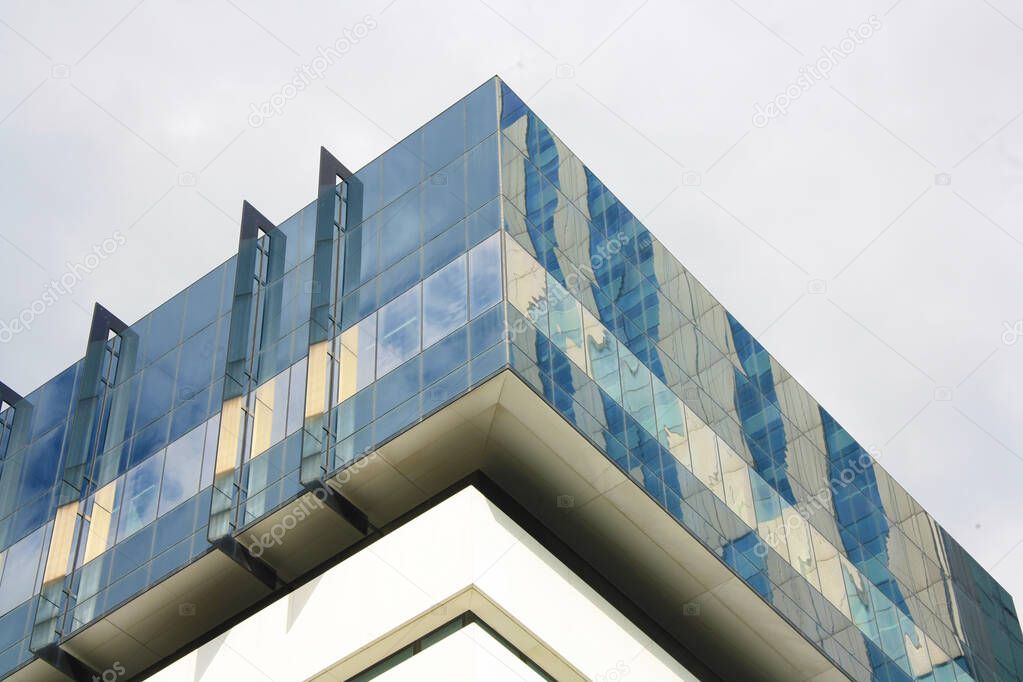 Detail view of the top of a modern building. Square form design. Structure with blue and white  glass windows and lines, with a cloudy sky in background. Low angle view. 