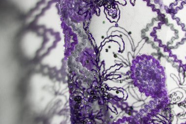 Şeffaf perdenin gölgesi ve duvardaki çiçek desenleri. Karmaşık işlemelerle süslenmiş mor kumaş. Tel, pullar ve boncuklar..