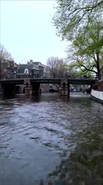 Amsterdam 'da bir kanal boyunca seyreden bir teknenin bakış açısı, Hollanda' daki geleneksel ev ve ağaçlarla çevrili park halindeki bisikletlerle tarihi bir taş köprüye yaklaşıyor..