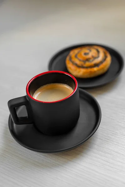 Sweet roll with poppy seeds and black cup of espresso on a white wooden table. Breakfast at home - black coffee and bun with poppy seeds. Selective focus.