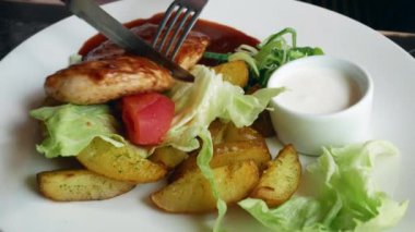 Sağlıklı öğle yemeği. Soslu ızgara tavuk, fırında patates ve beyaz tabakta taze sebze. Bıçakla tavuk eti kesmek..