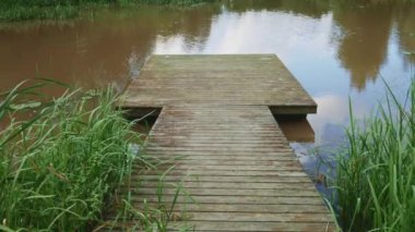 Sakin bir gölün kıyısındaki ahşap iskele bulutlu bir yaz gününde yeşil çimlerle çevrili. Bulutlar ve ağaçlar suya yansıyor..