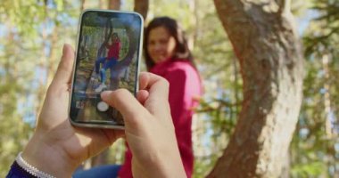 Akıllı telefon kullanan ve ağaçta oturan güzel, mutlu beyaz bir kadının resimlerini çeken ellerin yakın çekim görüntüleri..