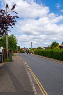 Norwich, UK- 17 Mayıs 2025: Norwich, Norfolk 'ta çalıları ve ağaçları olan sakin bir yerleşim yeri, mavi bulutlu bir yaz gökyüzünün altında tipik bir İngiliz banliyö yolunu gösteriyor.