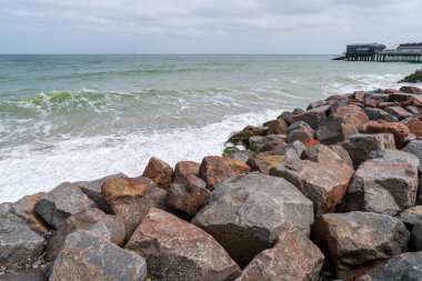 İngiltere 'nin Cromer rıhtımında büyük taşları ve okyanus dalgalarıyla Rocky kıyı savunma dalgaları, İngiliz Norfolk sahil beldesinde beyaz sörf ve Viktorya dönemi rıhtımı yapısına sahip deniz savunma kayaları